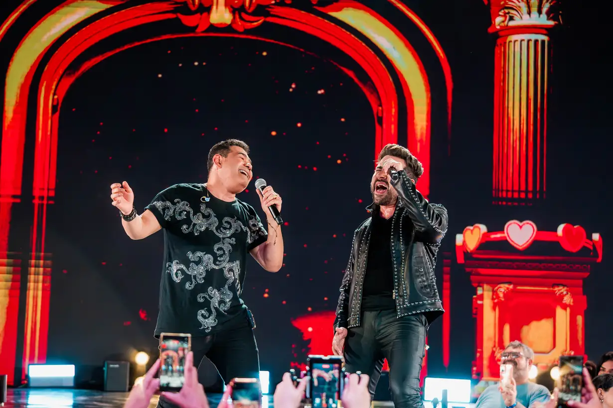 Batista Lima e Léo Lira cantam juntos no palco, diante de um arco luminoso vermelho.
