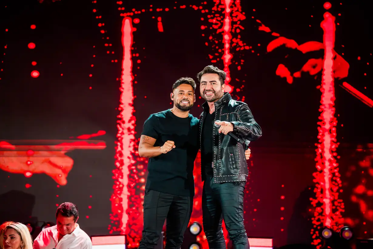 Dois cantores se abraçam no palco, sob luz vermelha.