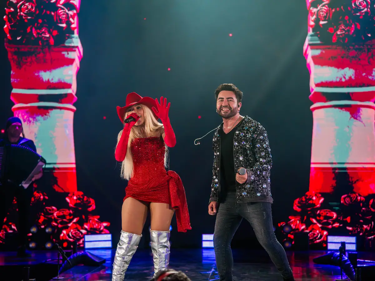 Músico com sanfona, cantora de chapéu e vestido vermelhos e cantor se apresentam no palco.